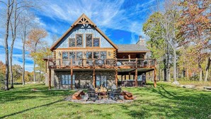A large log cabin with a stone foundation, a balcony, and a fire pit in the front yard. - Charming 6BR Chalet - HOT TUB - Mountain View (McHenry)