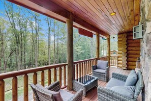 A log cabin with a wooden deck, wicker furniture, and a view of the forest. - Rustic 5BR Cabin - HOT TUB - Swanton (Swanton)