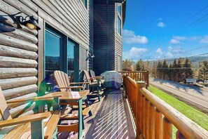 A wooden deck with chairs and a table, a covered grill, and a view of a road and trees.