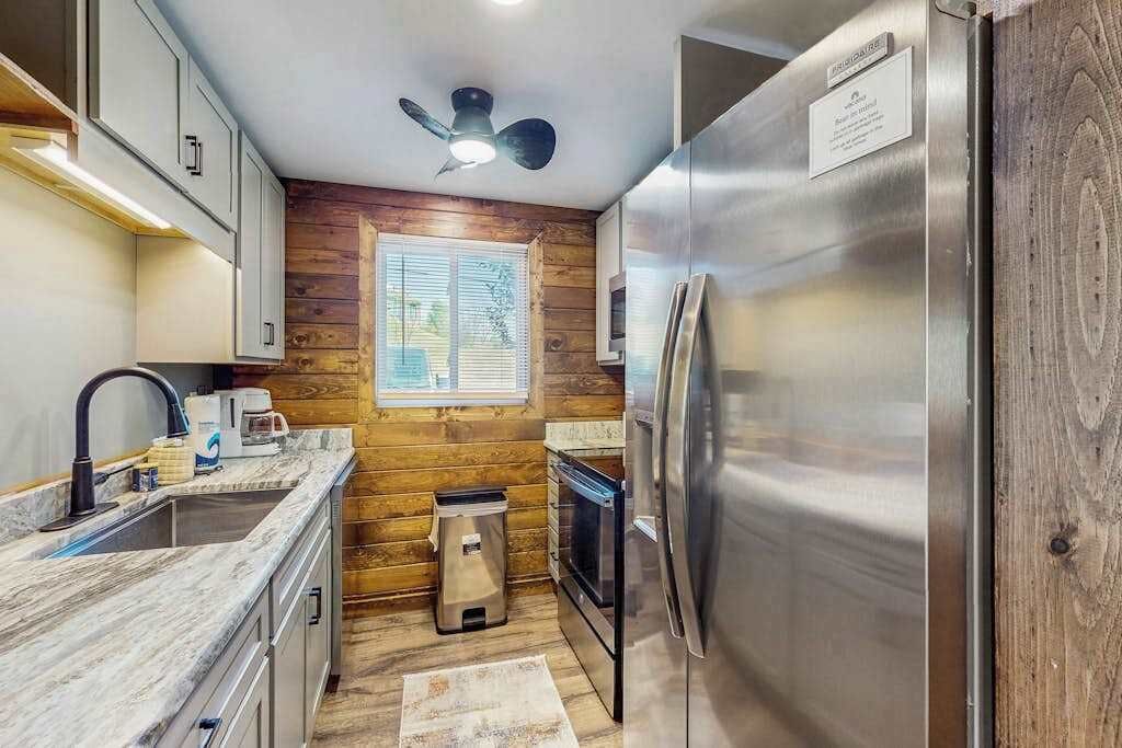 A compact kitchen with wooden walls, a stainless steel refrigerator, a sink, and a ceiling fan.