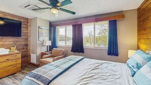 Un dormitorio con cama, una silla, un tocador, ventilador de techo y ventana con cortinas.