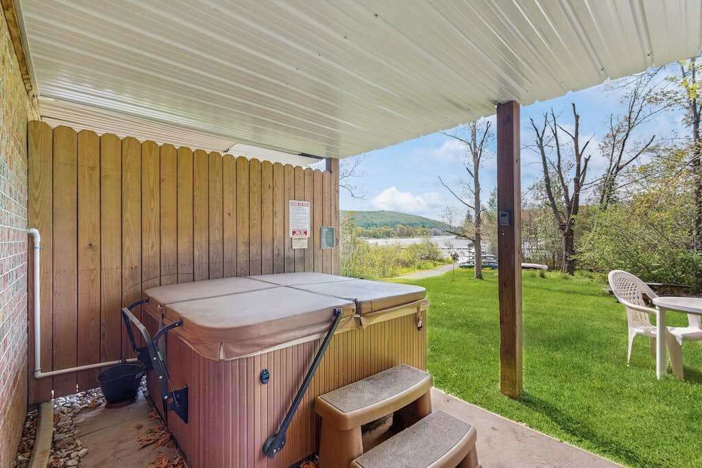Un jacuzzi bajo un porche cubierto, con valla de madera, zona de césped y vistas a colinas al fondo.