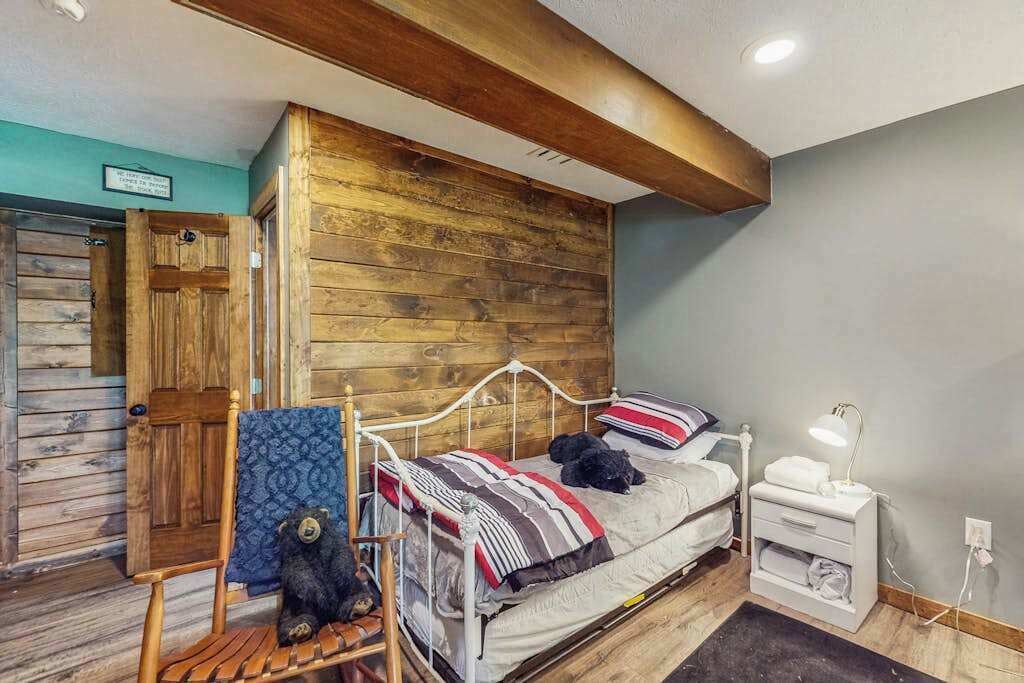 A bedroom with a wooden headboard, a bed with a striped blanket, a bedside table with a lamp, and a chair with a teddy bear.
