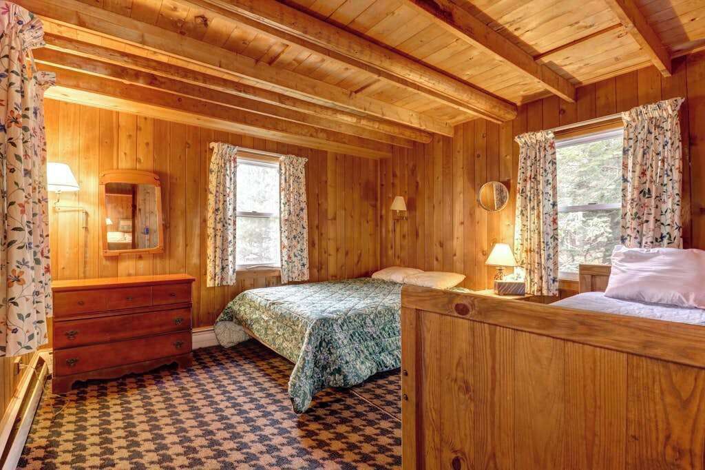 Dormitorio de cabaña de madera con piso de cuadros, una cama, un tocador y dos ventanas con cortinas.