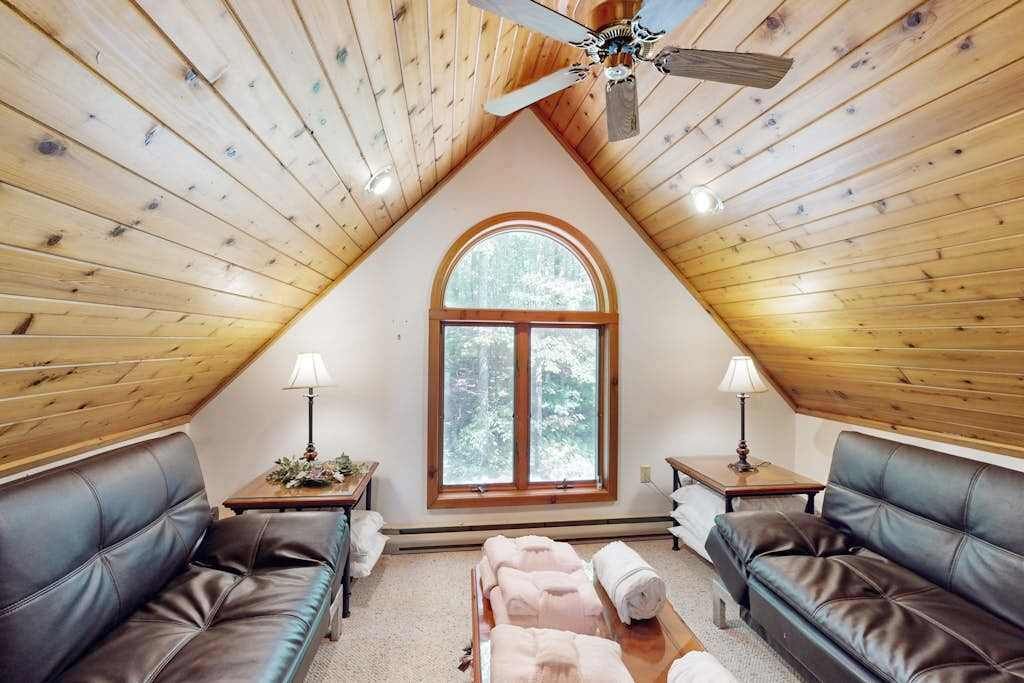 A cozy attic bedroom with wooden walls, a large arched window, two leather sofas, and a ceiling fan.