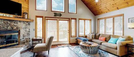 A spacious living room with a stone fireplace, wooden ceiling, large windows, and a cozy seating area.