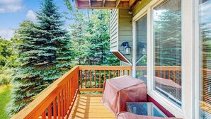 Une terrasse en bois dotée d’un espace salon couvert, d’une fenêtre avec des stores et d’une vue sur une forêt de conifères.