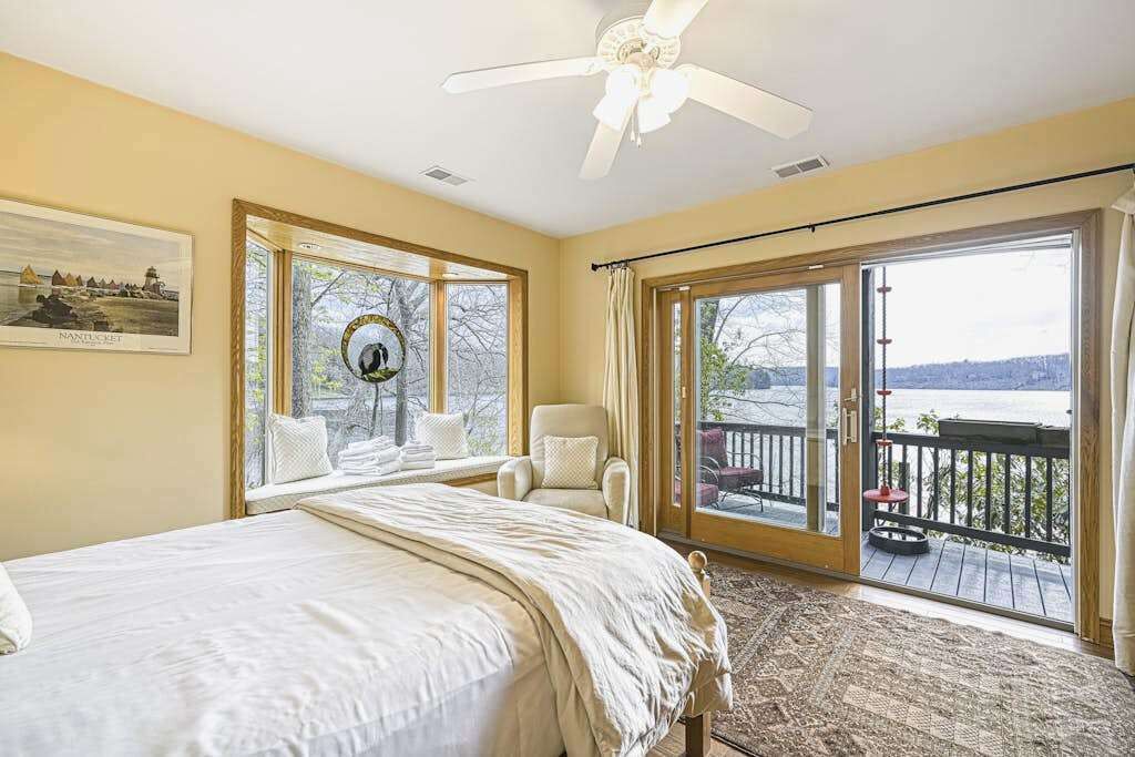 A bedroom with a bed, a chair, a ceiling fan, a large window, and a balcony with a view.