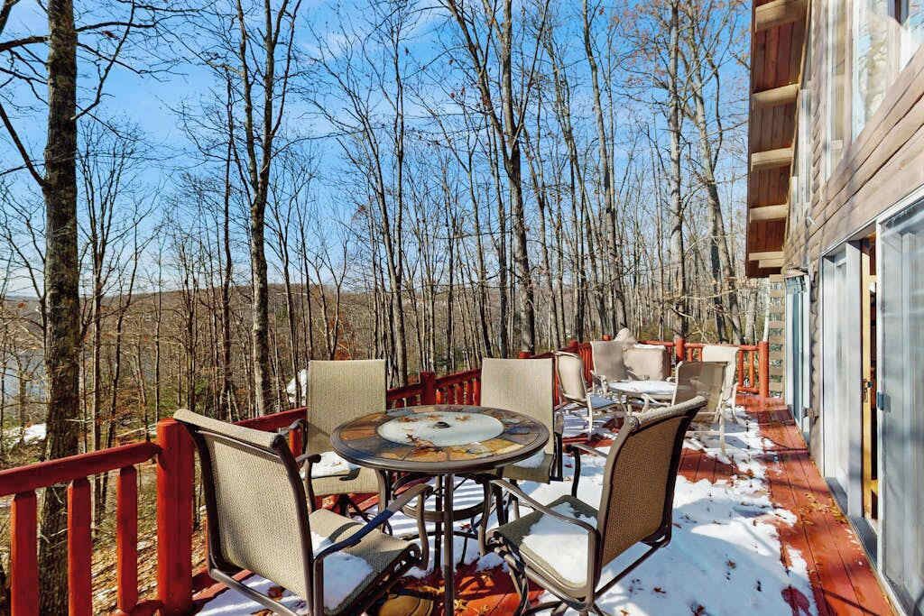 A covered outdoor patio with a round table and chairs, surrounded by a wooden deck and red railings, overlooking a forested area.