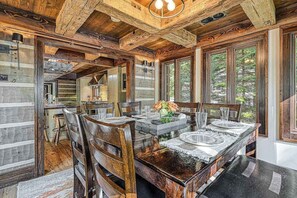 A dining area with a wooden table set for four, surrounded by chairs, with a view of trees through large windows. - Lakefront 7BR Chalet - HOT TUB - Swanton (Swanton)