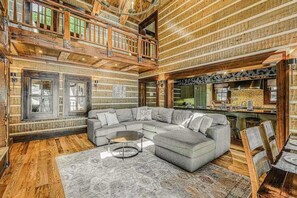 A log cabin interior with a large L-shaped sectional sofa, a dining table with chairs, and a kitchen area with a large island. - Lakefront 7BR Chalet - HOT TUB - Swanton (Swanton)