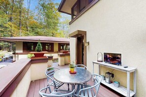 Een terras buiten met een ronde tafel en stoelen, een televisie op een standaard en een klein plankje met spullen.