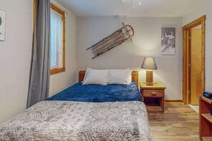 A bedroom with a bed, a bedside table with a lamp, a window with blinds, and a door. - 3BR - Lakeside Retreat - Pool (McHenry)