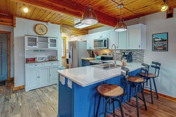 A kitchen with a central island, bar stools, white cabinets, a clock, and a sign that reads 'COME ON IN.'