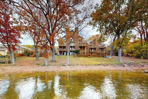 A multi-story building with a wooden deck, surrounded by trees with autumn foliage, and a body of water in the foreground. - Charming 3BR Condo - LAKE VIEW - McHenry (McHenry)