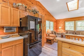 Interior de una cabaña de madera con cocina, comedor y sala de estar con sofá y lámpara.