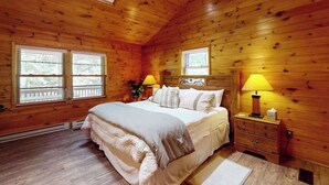 Quarto com painéis de madeira, uma cama, duas janelas, uma mesa de cabeceira e um abajur.