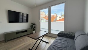 Uma sala de estar moderna com televisão de tela plana, uma mesa de madeira, um sofá, um vaso com planta e uma porta de vidro deslizante para o exterior.