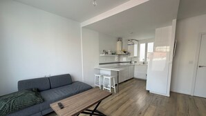 Um espaço de estar moderno com sofá cinzento, mesa de centro de madeira e área de cozinha com armários e bancada brancos.