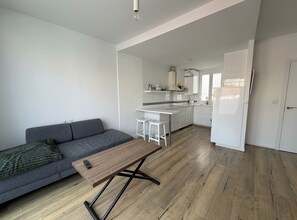 Un espacio habitable moderno con un sofá gris, una mesa de centro de madera y una cocina con armarios blancos y encimera blanca.