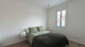 Quarto com cama, mesa de cabeceira, luminária, janela e vista para edifícios.