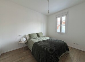 Un dormitorio con cama, mesita de noche, lámpara, ventana y vistas a edificios.