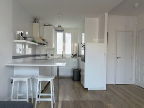 A modern kitchen with white cabinetry, a central island, two white bar stools, and a window providing natural light. - Apt - Sleeps 4 - Pet Friendly - Central Location (A Coruña)