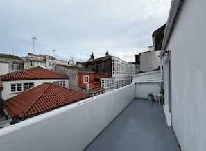 Una terraza en la azotea con vistas a edificios cercanos, una pared blanca y una barandilla de metal.