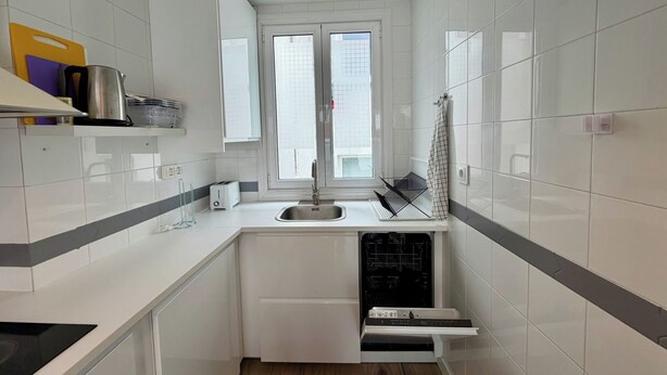 Uma cozinha compacta com armários brancos, máquina de lavar louça embutida e uma janela que traz luz natural.
