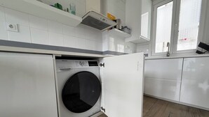Uma lavanderia compacta com máquina de lavar frontal, uma prateleira pequena com artigos de higiene pessoal e uma janela com vista.