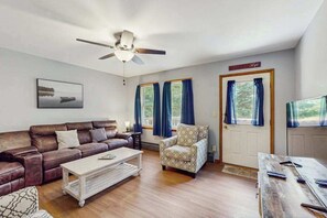 Ein Wohnzimmer mit braunem Ledersofa, einem gemusterten Sessel, einem weißen Couchtisch und einem Holzessttisch mit Fernbedienungen.