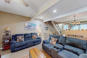 Ein Wohnzimmer mit zwei Sofas, einem Couchtisch, einem Bücherregal, einem Gemälde, einer Treppe und einer Küchenzeile.
