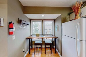 A compact kitchen with a dining table, two chairs, a white refrigerator, and a fire extinguisher on the wall.