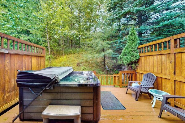 A hot tub with a cover, a small bench, and two Adirondack chairs on a wooden deck surrounded by a wooden fence and trees.