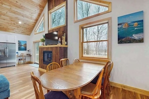 A dining area with a wooden table and chairs, a fireplace, and large windows with a view of trees. - Modern 4BR Cabin - Movie Theater - HOT TUB (McHenry)