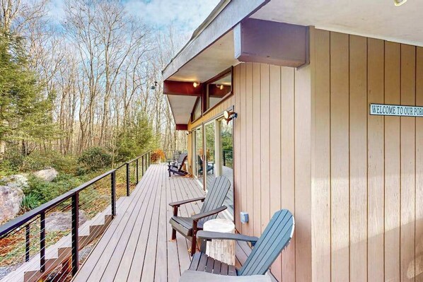 A wooden deck with Adirondack chairs, a sign reading 'WELCOME TO OUR PORT,' and a forested background.