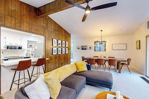 Una sala de estar moderna con un sofá seccional gris, un muro decorativo de madera, un comedor con una mesa y sillas de madera, y una cocina con armarios blancos y una barra.