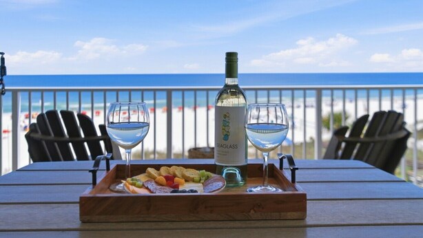 Una bandeja de madera con fruta y dos copas de vino sobre una mesa con vista a la playa.