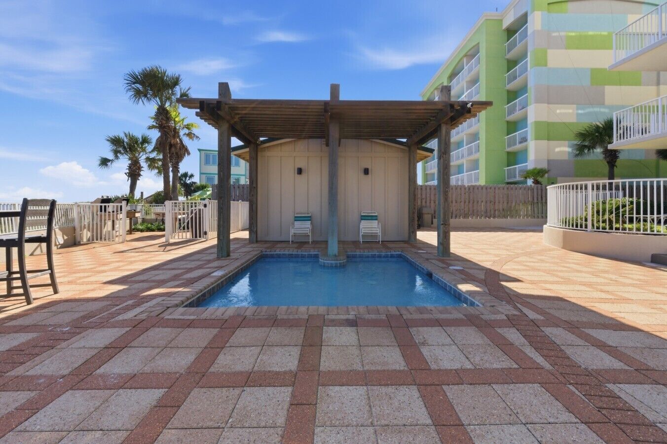Una piscina con un pérgola de madera, sillas de descanso y un edificio de varios pisos al fondo.