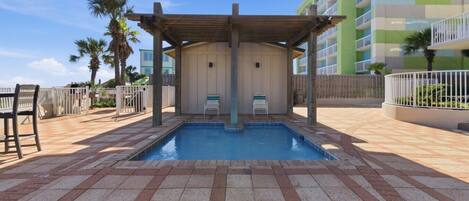 Una piscina con un pérgola de madera, sillas de descanso y un edificio de varios pisos al fondo.