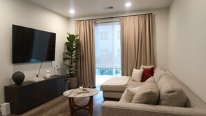 Een moderne woonkamer met een flatscreen televisie, een donker houten dressoir, een ronde salontafel, een beige bank en een groot raam met gordijnen.