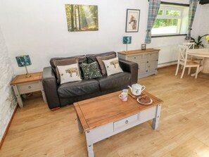 A cozy living room with a brown leather sofa, a wooden coffee table, and a dining area with a white table and chairs. - Daffodil Cottage (Treorchy)