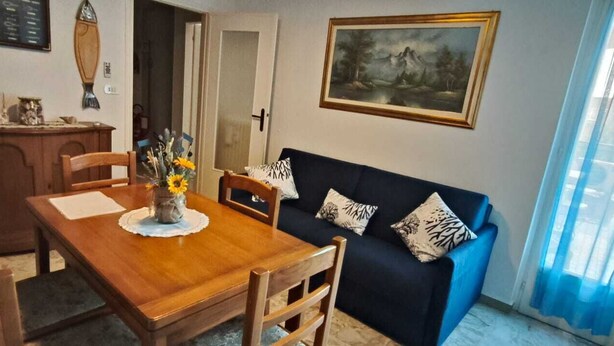 Uma área de refeições com mesa e cadeiras de madeira, um sofá azul escuro com almofadas estampadas, um vaso com flores e um quadro de paisagem na parede.