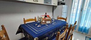 Ein Esstisch mit blauer Tischdecke, Holzstühle, einer Weinflasche und einer mit 'HOME' beschrifteten Kiste in einem Raum mit weißen Schränken und einem fenster mit Vorhängen.