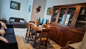 Uma sala de estar com um sofá escuro, uma mesa de jantar de madeira com cadeiras, um armário com portas de vidro e uma televisão.