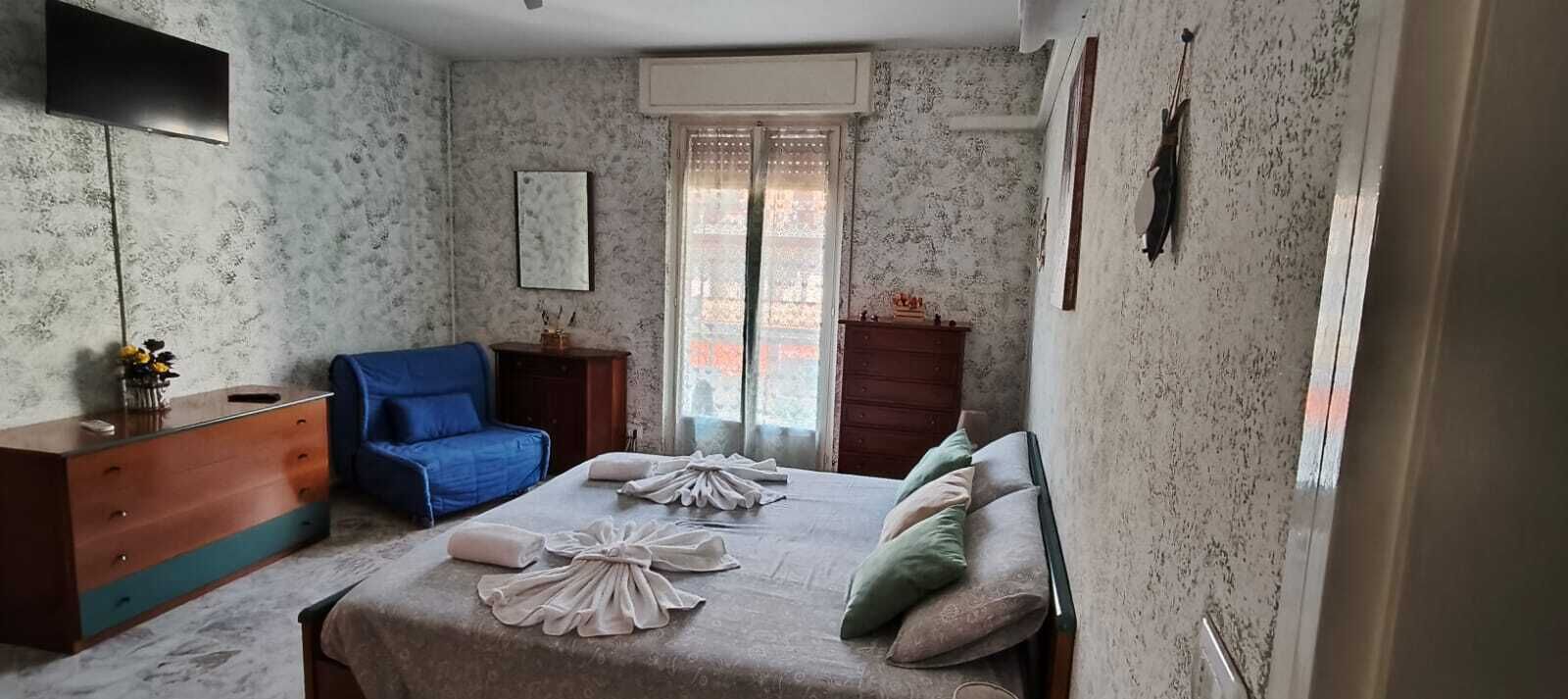 Een slaapkamer met een bed, een blauwe stoel, een dressoir, een televisie en een raam met gordijnen.
