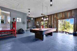 Une table de billard dans une pièce aux murs en bois, avec une échelle rouge et une grande fenêtre donnant sur la forêt.