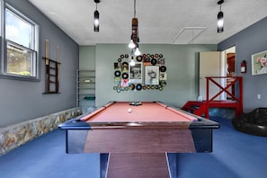 A pool table with a red felt surface, surrounded by a blue carpeted floor, with a wall decorated with vinyl records and framed pictures.