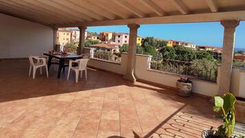 Una terrazza coperta con tavolo e sedie, un vaso di fiori e vista sul mare e sugli edifici.