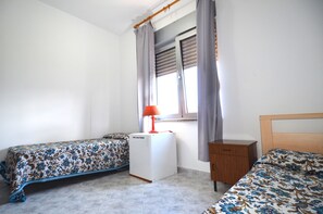 Ein Schlafzimmer mit zwei Einzelbetten, einer kleinen weißen Heizung, einem Holznachttisch und einem Fenster mit Vorhängen.